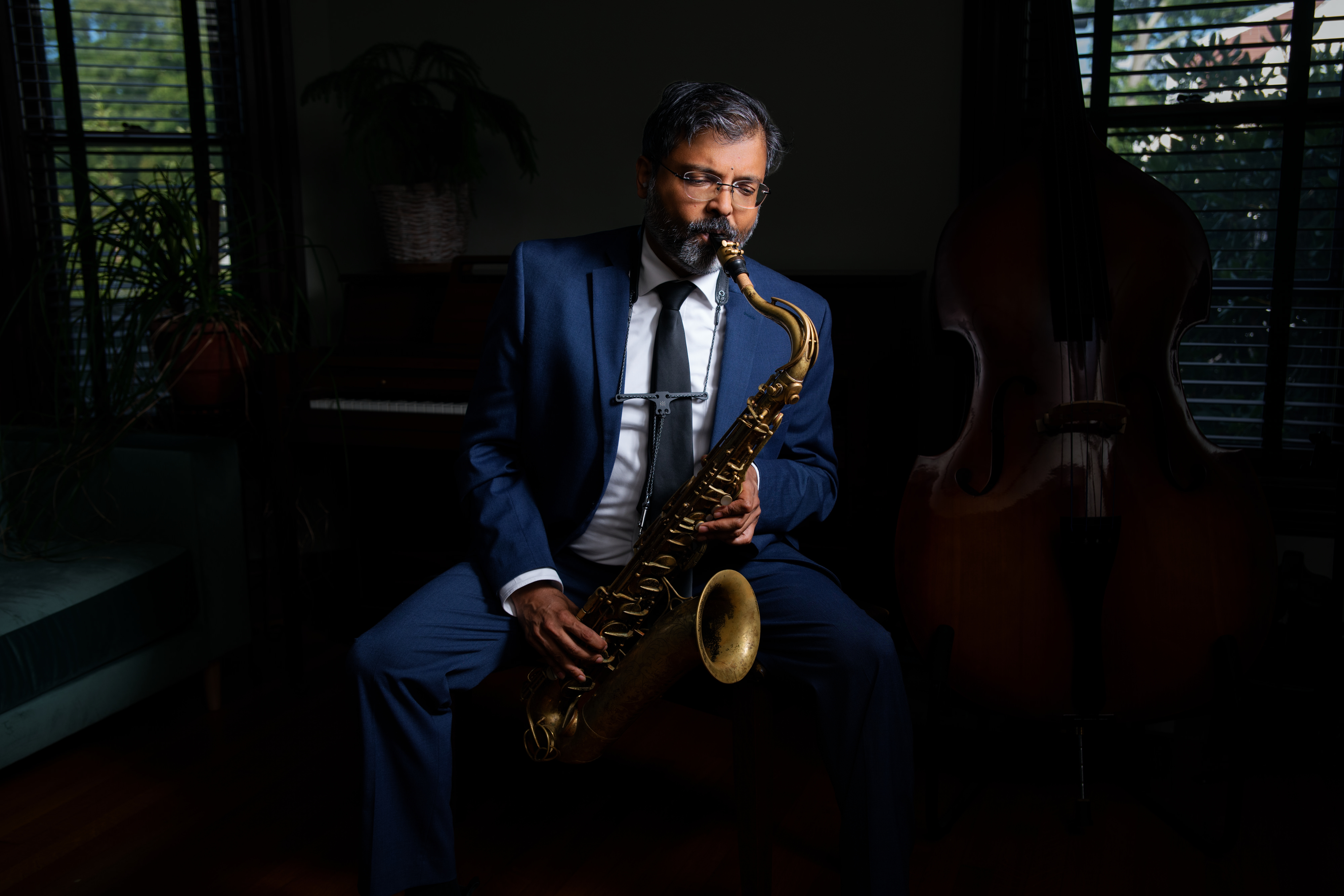 Sharat Somashekara in a blue suit playing tenor saxophone beside a piano and upright bass